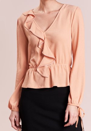 Perzikkleurige blouse met lange mouwen, met ruches langs de V-hals en een tailleband, gecombineerd met een zwarte rok.