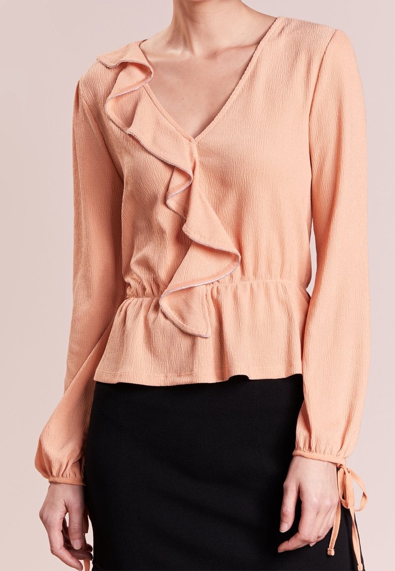 Blouse pêche à manches longues avec détail volanté le long du col en V et ceinture à nouer à la taille, associée à une jupe noire.