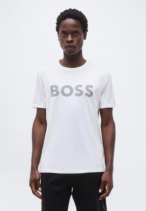 Joven con cabello rizado corto, vistiendo una camiseta blanca con la palabra "BOSS" en líneas grises en negrita, de pie contra un fondo claro y liso.