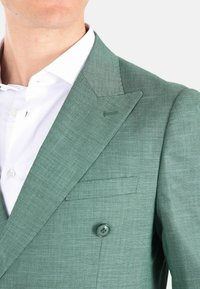 Blazer verde chiaro con rever a punta, tessuto testurizzato, due bottoni e una tasca sul petto; abbinato a una camicia bianca.