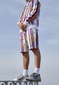 Gestreepte hoodie en bijpassende shorts in wit, rood, blauw en geel met smiley-gezicht prints; gecombineerd met grijze sportschoenen en witte sokken.