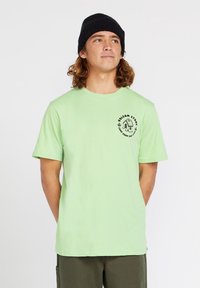 Camiseta de algodón de manga corta en verde claro, con cuello redondo y un pequeño logotipo gráfico negro en el lado izquierdo del pecho.