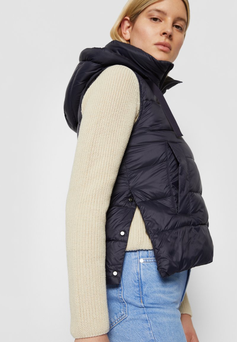 Marineblaues Steppweste mit Kapuze, seitlichen Druckknöpfen und vorderen Taschen, getragen über einem cremefarbenen Strickpullover. Hat ein verkürztes Design.