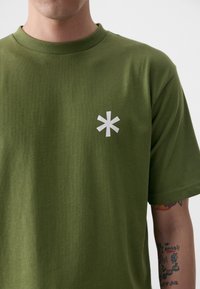 Oliven grøn bomulds T-shirt med korte ærmer og rund halsudskæring. Har et hvidt asterisk grafisk motiv på venstre brystområde.