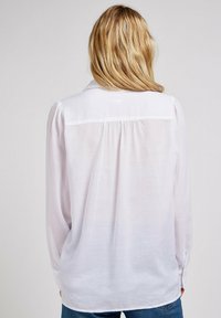 Blusa bianca con una vestibilità rilassata, maniche lunghe e polsini con bottone. Tessuto leggero con una sottile lucentezza e un dettaglio leggermente trasparente nella parte posteriore.
