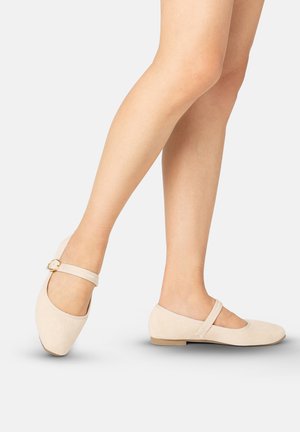 Legs wearing beige Mary Jane flats with a thin strap and gold buckle on a light background.