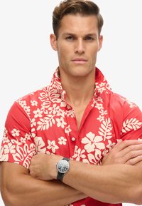 Camisa corta de manga larga roja con estampado de flores y hojas blancas de hibisco. El modelo lleva un reloj de muñeca negro con caja plateada.