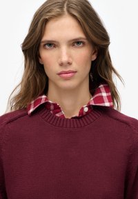 Burgundy knit sweater with a ribbed neck, layered over a red and white checkered shirt, featuring a buttoned collar and visible stitching details.