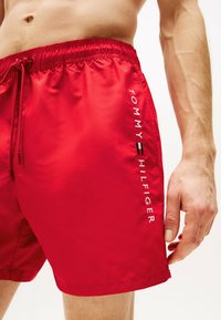 Røde svømmedragter lavet af glat stof med elastisk taljebånd. Har en hvid "TOMMY HILFIGER" logo trykt lodret på siden.