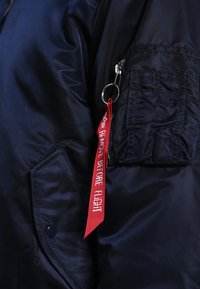 Jūrinė mėlyna bombonešio striukė su lygiu, blizgiu paviršiumi. Keturios užtrauktuko kišenės rankovėje, kurios akcentuotos raudonu užrašu "PAŠALINTI PRIEŠ SKRYDĮ".
