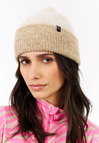 Woman wearing a beige knit beanie and a pink and beige striped zip-up jacket, with long dark hair and natural makeup.