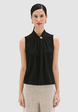 Donna con capelli pettinati all'indietro che indossa orecchini a cerchio dorati, un top nero senza maniche con una spilla decorativa e pantaloni chiari e strutturati.