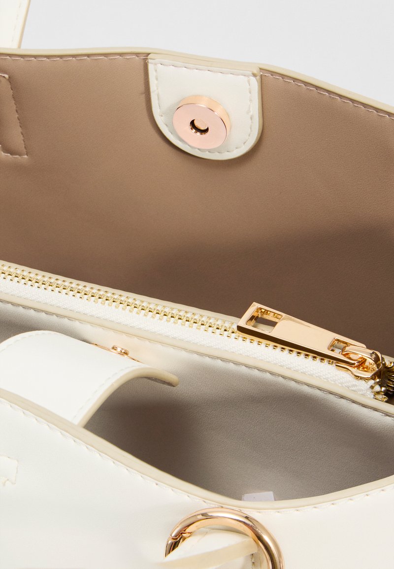 Gros plan de l'intérieur d'un sac à main beige et blanc montrant une fermeture éclair dorée, un fermoir magnétique, des coutures et un détail en métal rond.