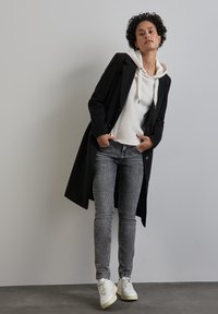 Manteau noir long sur un hoodie blanc, jeans gris et baskets blanches ; tissu lisse, design structuré et coupe décontractée ; le mannequin pose les mains dans les poches.