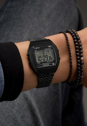Montre numérique noire avec un boîtier rectangulaire, des accents argentés et un bracelet en maille. L'écran affiche l'heure, la date et les fonctions de chronomètre.