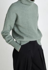 Hellgrüner Rollkragenpullover aus weichem Material mit lockerer Passform, ausgestattet mit gerippten Bündchen und Saum, kombiniert mit hoch taillierten schwarzen Hosen.
