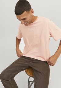 Lichtroze katoenen t-shirt met korte mouwen en een ronde hals, voorzien van "JACK & JONES PREMIUM" in witte tekst. Gecombineerd met een paar bruine broeken.