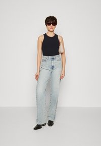 Top sin mangas negro con textura acanalada, jeans de tiro alto azules claros, tacones angulares negros, bolso pequeño beige, gafas de sol oscuras y grandes, fondo sencillo.