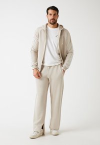 Beige sweatshirt en bijpassende broek van zacht, rekbaar materiaal. Met capuchon, voorzakken en zijsplitten op de broek. Witte sneakers.