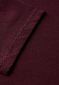 Bordeaux stofmonster met een gladde textuur, voorzien van een gevouwen mouw en subtiele stiksels langs de rand.