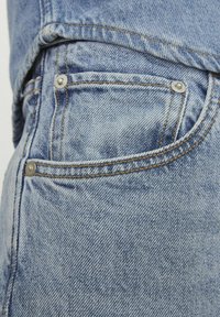 Svijetloplava traper tkanina sadrži džep sa srebrnim zakovicama, kontrastnim šavovima i blago teksturiranom površinom na prednjem dijelu hlača.