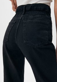Pantalon en denim noir à taille haute, avec une surface texturée, deux poches arrière et une taille froncée pour une coupe décontractée.