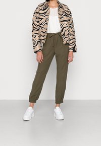 Veste courte rayée zèbre beige et noire, blouse blanche, pantalon vert olive retroussé et baskets blanches à plateforme. Les textures et motifs sont bien visibles.