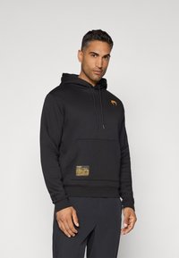 Black hoodie with kangaroo pocket, adjustable drawstring, orange logo on chest, and a gold patch on the pocket featuring brand details.