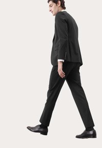 Schwarzer Anzug mit maßgeschneidertem Schnitt, bestehend aus einer Jacke und passenden Hosen. Trägt dazu dunkle Schuhe, die ein elegantes Design und eine glatte Textur zeigen.