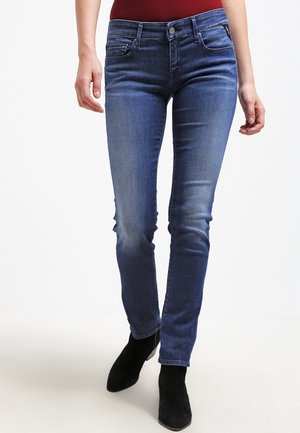 Femme portant un jean bleu ajusté et des bottines noires, debout devant un fond blanc uni.