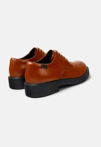 Zapatos de cuero marrón con un acabado suave, punta redonda y suela de goma negra gruesa. Presentan un diseño de cordones y una pequeña etiqueta de la marca en el talón.
