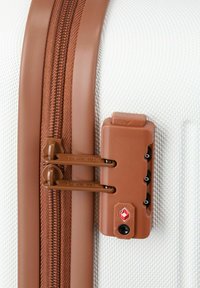 Valise rigide avec une surface texturée blanche, fermeture éclair marron et un cadenas intégré approuvé par la TSA. Les tirettes de la fermeture éclair sont également marron.