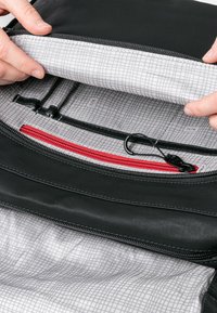 Schwarzes Innenfutter der Tasche mit einem hellgrauen, gittergemusterten Futter. Verfügt über eine rote Reißverschlusstasche, verschiedene Schlaufen und eine glatte Textur.