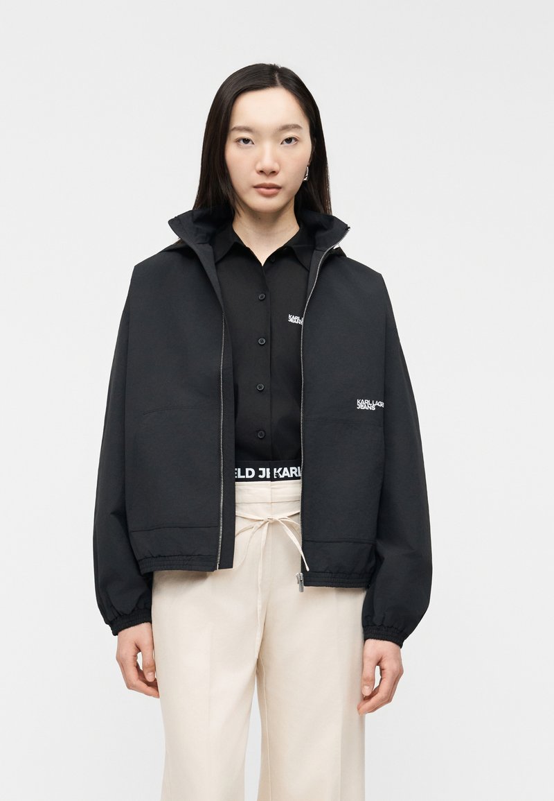 Γυναίκα με μακριά σκουρόχρωμα μαλλιά που φοράει μαύρο μπουφάν με φερμουάρ πάνω από μαύρο πουκάμισο με κουμπιά και ανοιχτό μπεζ παντελόνι, στέκεται μπροστά σε απλό φόντο.