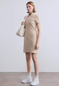 Μπεζ φόρεμα πόλο με κοντά μανίκια, κουμπιά στο γιακά, συνδυασμένο με λευκή τσάντα και αθλητικά παπούτσια. Λείο βαμβακερό ύφασμα, σχεδιασμένο μέχρι το γόνατο.