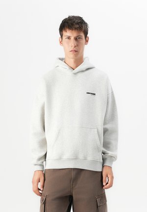 Jonge man in een lichtgrijze hoodie met een kangaroozak en een donker tekstlogo, gecombineerd met bruine cargobroek.