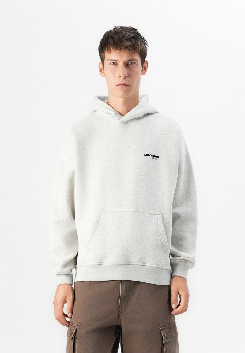 Młody mężczyzna w jasnoszarej bluzie z kapturem z przednią kieszenią i ciemnym logo, w połączeniu z brązowymi spodniami cargo.