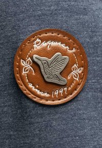 Brun läderlapp med en metallisk kolibri-emblemet, vit kursiv text som säger "Ragwear sedan 1997," broderad på en blå tygbakgrund.