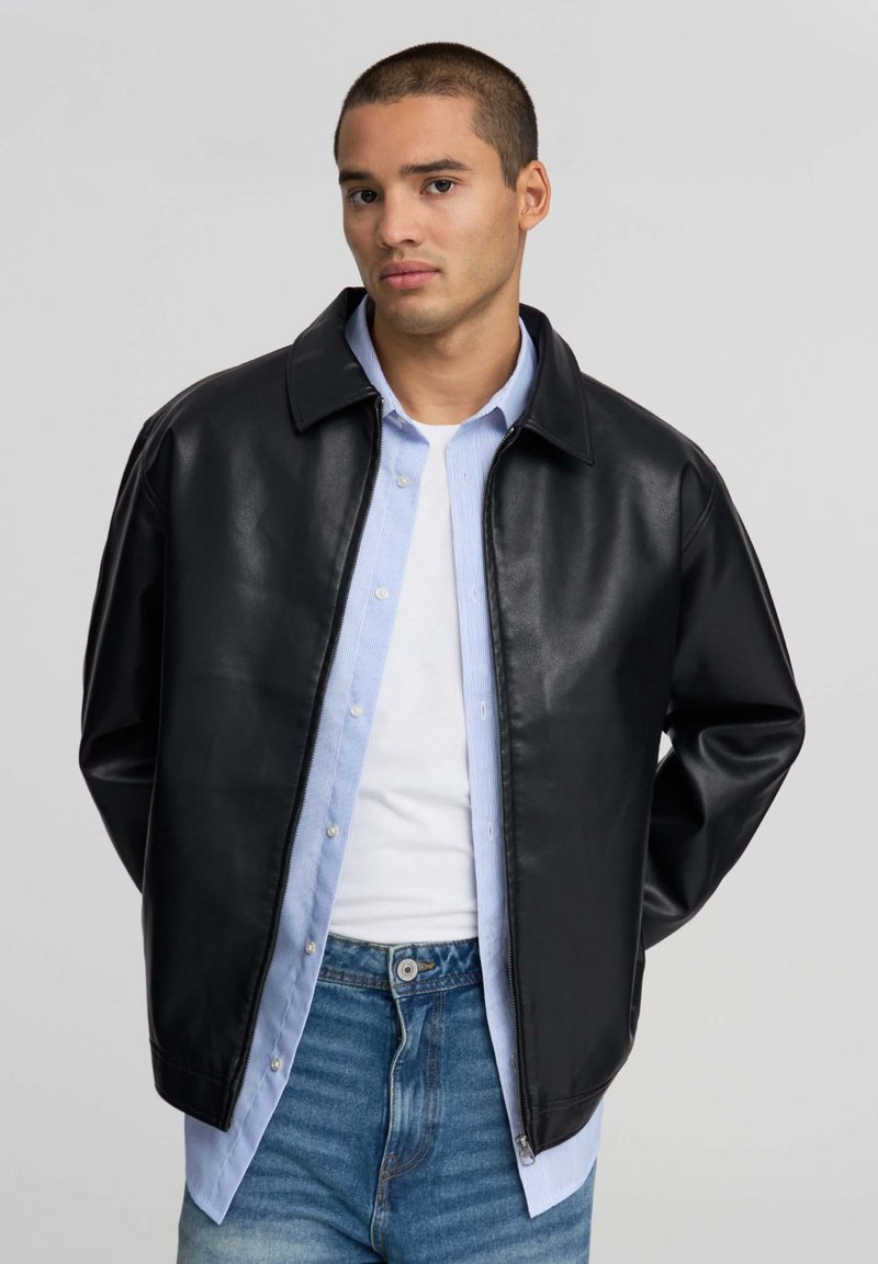 Giovane uomo che indossa una giacca di pelle nera sopra una camicia azzurra a maniche lunghe e una t-shirt bianca, abbinata a jeans blu, in piedi davanti a uno sfondo grigio.