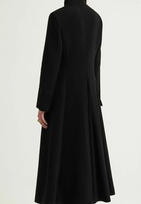 Cappotto lungo nero con colletto alto, vestibilità sartoriale e design posteriore plissettato. Realizzato in tessuto strutturato, con maniche lunghe e senza chiusure visibili.