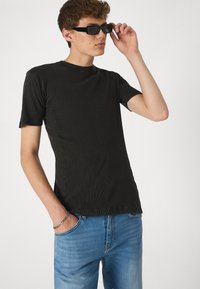 Crna rebrasta majica kratkih rukava u kombinaciji sa svijetloplavim trapericama. Model nosi crne naočale za sunce, ruka mu je u džepu, minimalistički dizajn.