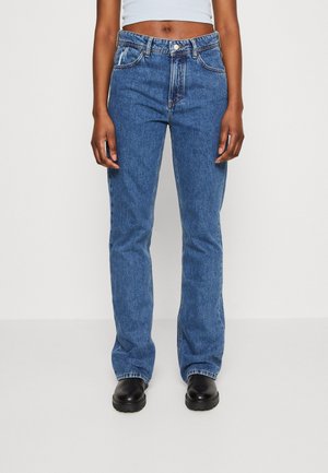 Personne portant un jean bleu taille haute à jambes droites et des chaussures noires, debout devant un fond clair uni, les bras détendus le long du corps.
