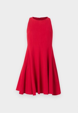 Frau steht, trägt ein ärmelloses rotes knielanges Kleid mit ausgestelltem Rock und weiße spitze flache Schuhe vor einem schlichten Hintergrund.