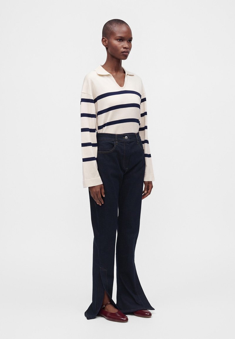 Personne portant un haut à manches longues rayé crème et marine, un pantalon foncé taille haute avec des fentes à l'avant, et des chaussures plates rouge foncé, debout sur fond blanc.