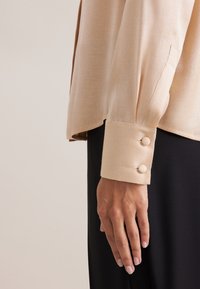 Blusa beige a maniche lunghe con polsini arrotolati e due bottoni rivestiti in tessuto, abbinata a un tessuto nero sottostante che mostra una consistenza liscia.