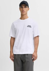 Valkoinen puuvillapaita, jossa on pyöreä kaula-aukko ja lyhyet hihat. Vasemmassa rinnassa mustalla brodeeratun "APPAREL STUDIOS" -logon.