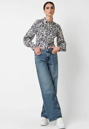 Frau, die mit Händen in den Taschen posiert, trägt ein gemustertes Hemd mit Knöpfen, weit geschnittene blaue Jeans und weiße Sneakers vor einem einfarbigen Hintergrund.