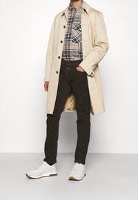 Manteau beige mi-long, chemise à carreaux boutonnée avec des tons foncés et clairs, pantalon marron foncé, baskets blanches avec des accents gris et des semelles marron.