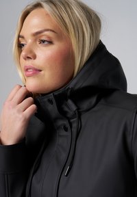 Veste imperméable noire avec un col haut, cordon de serrage et fermeture à pression, texture lisse et design ajusté, portée par une personne aux cheveux clairs.