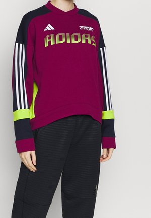 Person wearing a maroon Adidas sweatshirt with neon yellow and black accents and striped sleeves, paired with black textured pants.